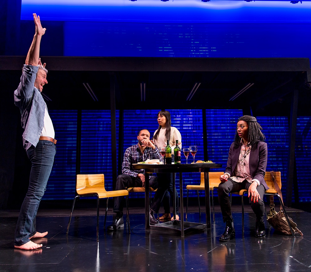 Barefoot man smiles and stretches upwards. Two women at the table stare unamused. Man at table stares outwards disassociated