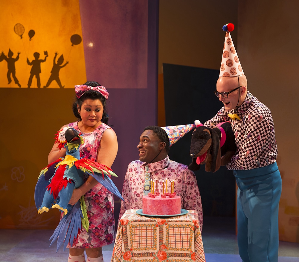 Woman on left holds a parrot puppet next to a man sitting in front of a cake while man on right holds a dachshund puppet