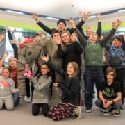 Children in a school library smile and raise their arms upward. An elephant prop and man in top hat appear at the center of the group.