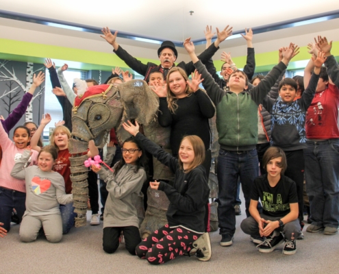 Children in a school library smile and raise their arms upward. An elephant prop and man in top hat appear at the center of the group.