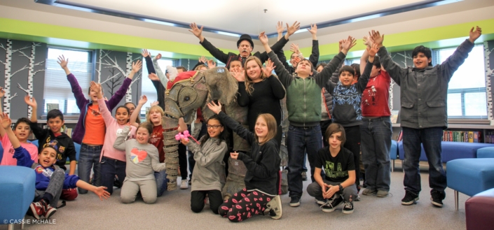 Children in a school library smile and raise their arms upward. An elephant prop and man in top hat appear at the center of the group.