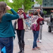 A group of patrons are outside on the sidewalk with their hands encircling their eyes to emulate binoculars while looking around