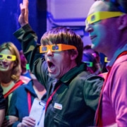 Audience members wearing bright-colored 3D glasses gasp in reaction to something out of frame
