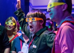 Audience members wearing bright-colored 3D glasses gasp in reaction to something out of frame