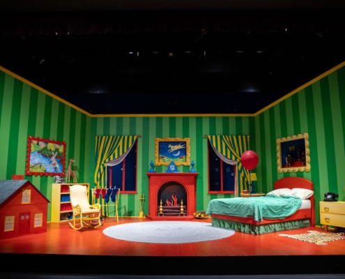 Colorful theatrical stage set designed as a whimsical children's bedroom, featuring green striped walls, bright orange-red floor, playful furniture, and decorations inspired by the book "Goodnight Moon," including a toy house, rocking chair, multicolored bookshelf, red fireplace with a flying cow picture, windows showing a starry night sky, and a green bed with a red balloon.