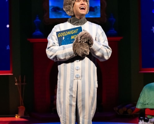 A person dressed in a gray bunny costume with tall ears and wearing blue-and-white striped pajamas stands on a stage holding a book titled “Goodnight Moon.” The background features a cozy room set with a red fireplace, blue walls decorated with stars, and a round rug on the floor. Plush toys and props are visible near the fireplace and on the floor.