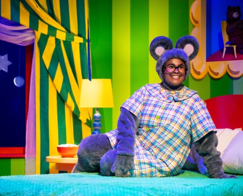 A person in a gray mouse costume with large round ears is lying on a green bed in a brightly colored room. The costume includes a plaid dress in shades of blue, yellow, and red. The background features green and yellow striped walls, a red bed frame, a white pillow, a yellow lamp on a nightstand, and a window with star decorations and striped curtains.