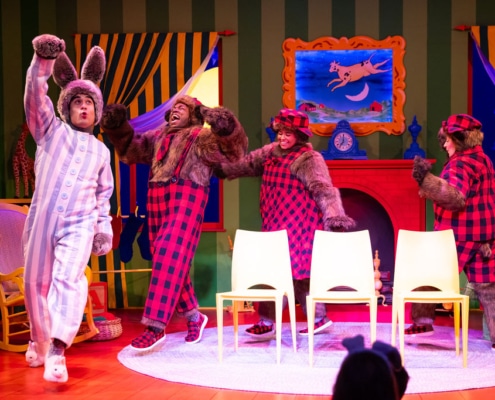 Four performers in animal costumes are on a colorful stage set resembling a cozy room. On the left, a performer dressed as a bunny in blue-and-white striped pajamas is stepping forward with one arm raised. To the right, three performers dressed as bears in furry costumes with red-and-black plaid outfits and matching hats are positioned near a red fireplace with a blue painting above it. In front of them are three white chairs arranged in a row. The background features green striped walls, windows with starry night scenes and striped curtains, and a rocking chair on the left side.