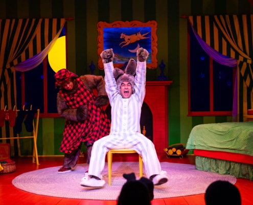 Two performers in animal costumes are on a colorful stage set designed like a cozy bedroom. In the foreground, a performer dressed as a bunny in blue-and-white striped pajamas is sitting on a yellow chair with both arms raised high. Behind the bunny, a performer dressed as a bear in a furry costume with a red-and-black plaid outfit and matching hat stands near a red fireplace. The background includes green striped walls, windows with starry night scenes and striped curtains, a green bed with a red frame, a yellow lamp on a nightstand, and a rocking chair on the left side.