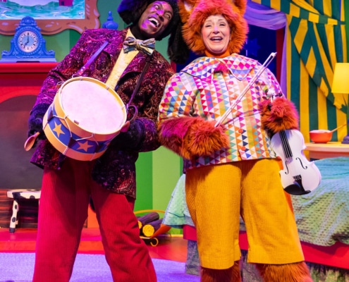 Two performers in animal costumes stand on a vibrant stage set resembling a cozy bedroom. The performer on the left is dressed as a dog, wearing a dark textured jacket with a large bow tie and bright red pants, and holding a round drum decorated with blue and yellow stars. The performer on the right is dressed as a cat in an orange furry costume, wearing a multicolored patterned shirt and yellow pants, and holding a white violin with a bow. The background includes green striped walls, a red fireplace with a blue painting above it, a yellow lamp on a nightstand, and striped curtains framing a window with a crescent moon and stars.
