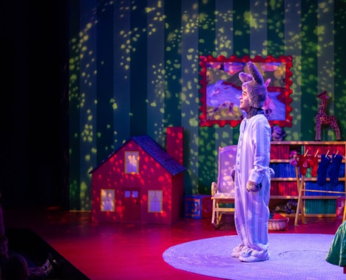 A performer dressed as a bunny stands on a colorful stage set designed like a cozy bedroom. The bunny costume includes blue-and-white striped pajamas and large floppy ears. The performer is standing on a circular rug with a green bed partially visible on the right. Behind them is a green striped wall decorated with a framed picture, a red toy house, a giraffe figurine, and a small shelf with hanging clothes. Warm, glowing lights create a starry pattern across the wall, adding a dreamy atmosphere to the scene.