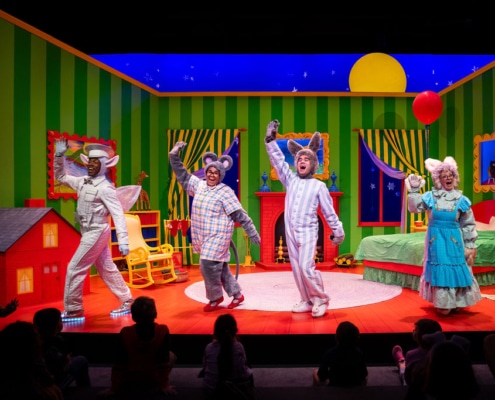 Four performers in colorful costumes stand on a vibrant stage set designed like a cozy bedroom. From left to right: the Tooth Fairy in a shiny silver outfit with a star belt and glowing shoes; a gray mouse wearing a plaid nightshirt; a bunny in blue-and-white striped pajamas; and a Grandma Bunny in a long turquoise dress with a white bonnet and apron. A red balloon is visible in the background near the bed. The set features green striped walls, a red bed with a green cover, a red toy house, a yellow rocking chair, windows showing a night sky with stars and a crescent moon, and a circular rug in the center. Children are seated in the foreground watching the performance.