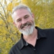 Man with white hair, mustache and beard wearing a black polo shirt and standing in front of a fall tree.