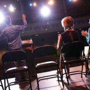 An image from the back of stage toward the seating area that features folding chairs where 2 people are seated and one individual is standing with arms outstretched as if he is auditioning.