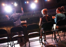 An image from the back of stage toward the seating area that features folding chairs where 2 people are seated and one individual is standing with arms outstretched as if he is auditioning.