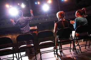 An image from the back of stage toward the seating area that features folding chairs where 2 people are seated and one individual is standing with arms outstretched as if he is auditioning.