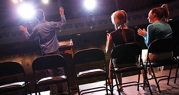 An image from the back of stage toward the seating area that features folding chairs where 2 people are seated and one individual is standing with arms outstretched as if he is auditioning.
