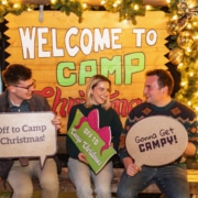 Three young people sit on a bench in front of a sign that reads Welcome to Camp Christmas. They are holding signs that read Off to Camp Christmas! and Gonna Get Campy!