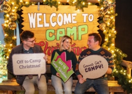 Three young people sit on a bench in front of a sign that reads Welcome to Camp Christmas. They are holding signs that read Off to Camp Christmas! and Gonna Get Campy!