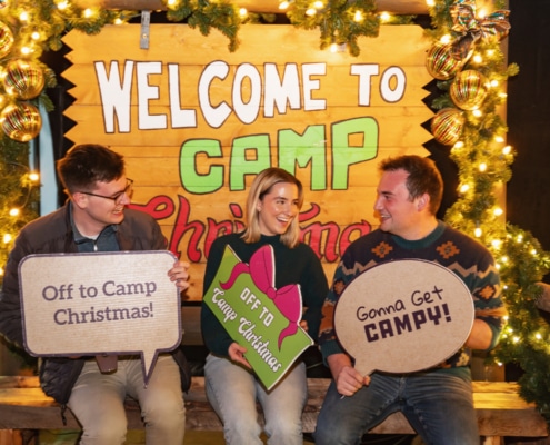 Three young people sit on a bench in front of a sign that reads Welcome to Camp Christmas. They are holding signs that read Off to Camp Christmas! and Gonna Get Campy!