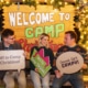 Three young people sit on a bench in front of a sign that reads Welcome to Camp Christmas. They are holding signs that read Off to Camp Christmas! and Gonna Get Campy!