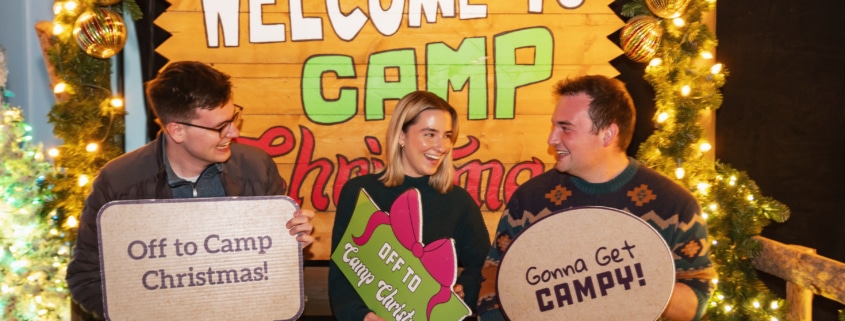 Three young people sit on a bench in front of a sign that reads Welcome to Camp Christmas. They are holding signs that read Off to Camp Christmas! and Gonna Get Campy!