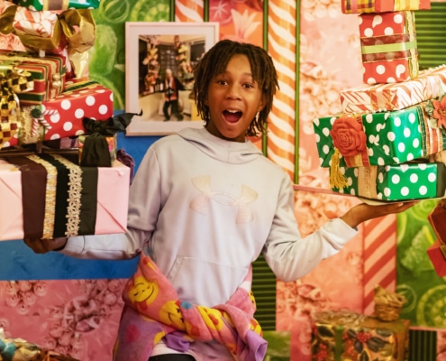 A young boy with a smiley face sweatshirt tied around his waist balances two stacks of presents in his hands.