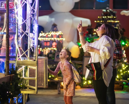 A young girl wearing a pink dress looks up in awe as her mother points up at something off camera. There are Christmas decorations, toys, and lights all around them.