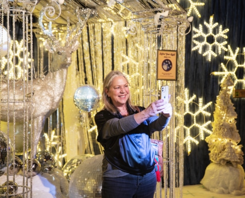 A woman with blonde hair and wearing a black top takes a selfie in front of a reindeer statue that is covered in silver glitter. There are light up snowflakes, strands of crystals, and disco balls in the background.