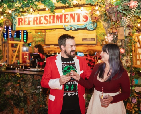 A couple wearing holiday attire smiles at each other and makes a cheers motion with their drinks. They are standing in front of a refreshment stand that is decorated with lights and garland.