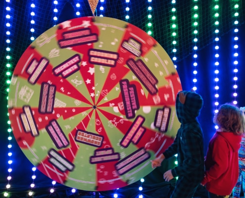 Three young children spin a large wheel. The wheel is green and red and lists out Holiday Superpowers.