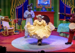 A brightly colored bedroom done in primary colors with an actor dressed as a bunny and an actress in the foreground dressed a furry animal curtsying in a yellow dress.
