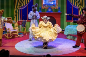 A brightly colored bedroom done in primary colors with an actor dressed as a bunny and an actress in the foreground dressed a furry animal curtsying in a yellow dress.
