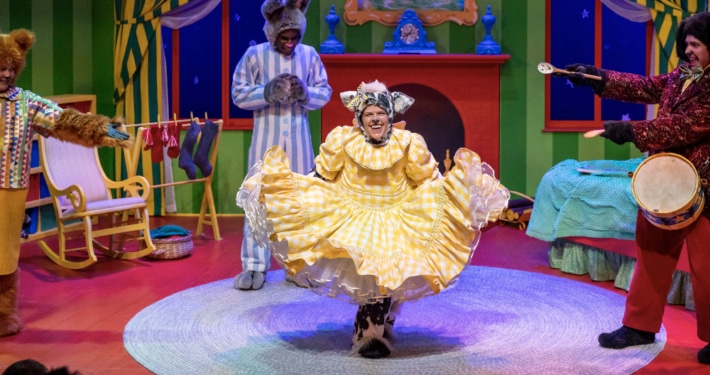 A brightly colored bedroom done in primary colors with an actor dressed as a bunny and an actress in the foreground dressed a furry animal curtsying in a yellow dress.