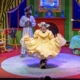 A brightly colored bedroom done in primary colors with an actor dressed as a bunny and an actress in the foreground dressed a furry animal curtsying in a yellow dress.