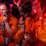 A group of individuals dressed in orange space suits talk anxiously on phones