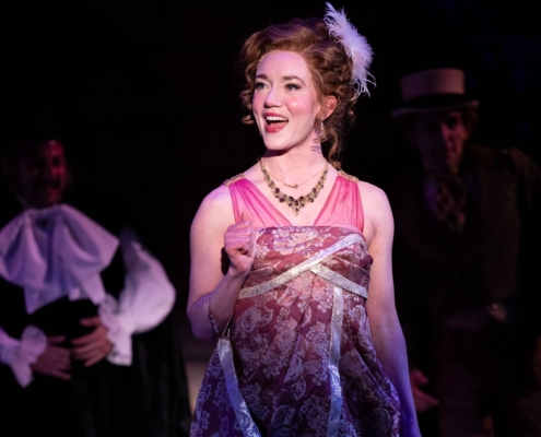 A woman in a pink patterned regency dress, smiles and speaks to the audience.