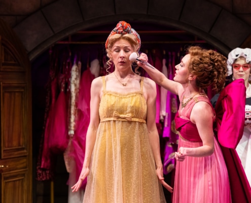 A woman in a pink regency dress with a large pink bow puts makeup on a woman in a yellow regency dress.