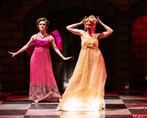 A woman in a pink regency dress with a large pink bow and a woman in a yellow regency dress dance across the stage.