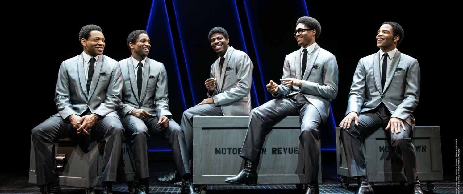 Five men in gray suits sit on top of boxes on a stage.