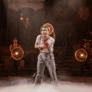 A performer stands center stage offering a red flower, with bright round lamps glowing behind and low fog drifting across the floor.