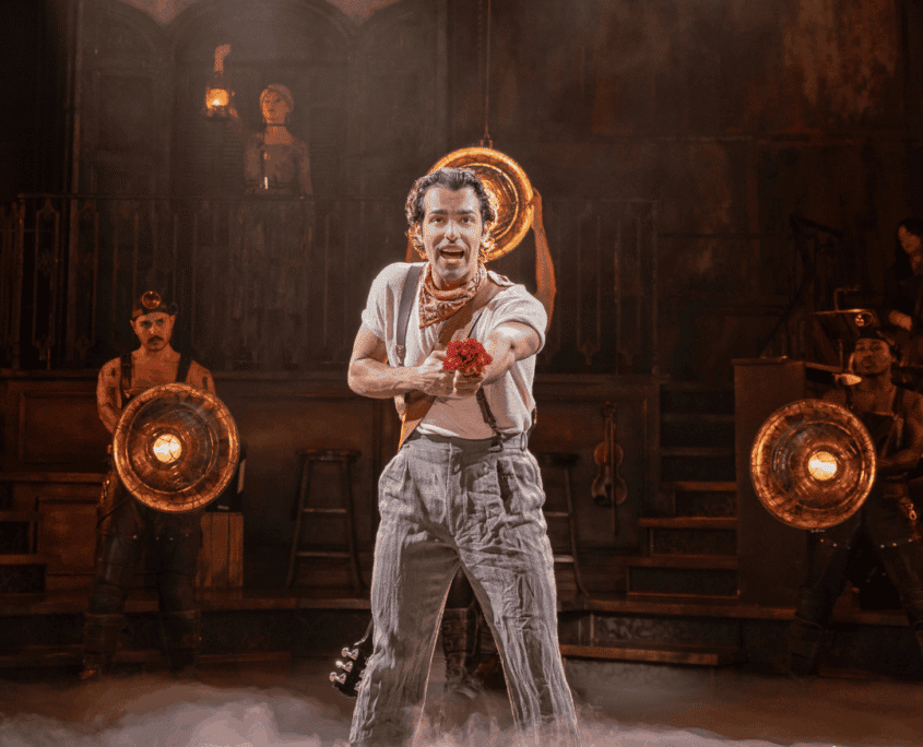 Credit: Jose Contreras (Orpheus) in Hadestown North American Tour, 2026. Photo by Evan Zimmerman for MurphyMade. A performer stands center stage offering a red flower, with bright round lamps glowing behind and low fog drifting across the floor.