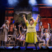 A central performer raises two bottles high in the air while a cheering group gathers behind on a colorful, celebratory stage set.