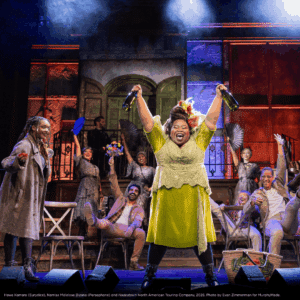 Credit: Hawa Kamara (Eurydice), Namisa Mdlalose Bizana (Persephone) and Hadestown North American Touring Company, 2026. Photo by Evan Zimmerman for MurphyMade. A central performer raises two bottles high in the air while a cheering group gathers behind on a colorful, celebratory stage set.
