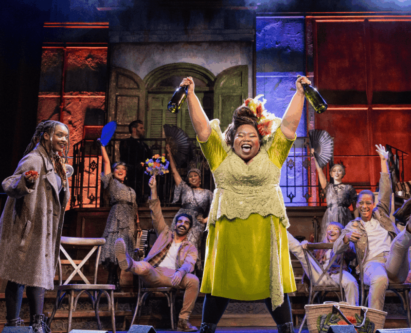 Credit: Hawa Kamara (Eurydice), Namisa Mdlalose Bizana (Persephone) and Hadestown North American Touring Company, 2026. Photo by Evan Zimmerman for MurphyMade. A central performer raises two bottles high in the air while a cheering group gathers behind on a colorful, celebratory stage set.