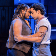 Two people stand very close, holding each other with foreheads touching under cool blue stage lighting.