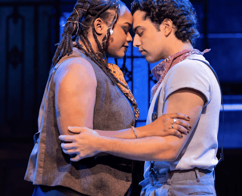 Credit: Hawa Kamara (Eurydice) and Jose Contreras (Orpheus) in Hadestown North American Tour, 2026. Photo by Evan Zimmerman for MurphyMade. Two people stand very close, holding each other with foreheads touching under cool blue stage lighting.