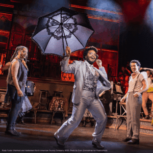 Credit: Rudy Foster (Hermes) and the Hadestown North American Touring Company, 2026. Photo by Evan Zimmerman for MurphyMade. A performer dances forward holding an open black umbrella, surrounded by other performers on a warmly lit stage.