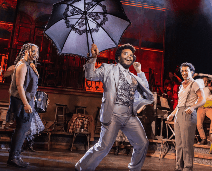 Credit: Rudy Foster (Hermes) and the Hadestown North American Touring Company, 2026. Photo by Evan Zimmerman for MurphyMade. A performer dances forward holding an open black umbrella, surrounded by other performers on a warmly lit stage.
