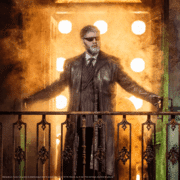 A person in a long dark coat stands on a balcony with arms spread wide, silhouetted by intense golden lights and swirling smoke.